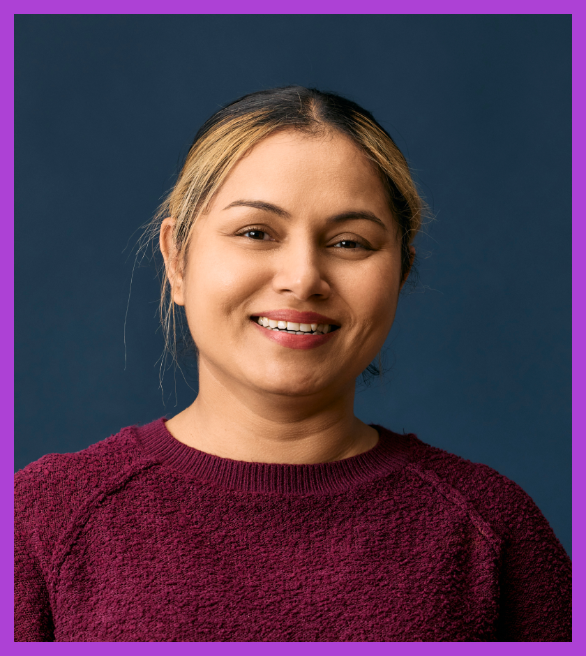 Camy is a middle-aged Indian woman with light brown skin and dark hair and dark eyes. She is wearing a maron jumper and smiling at the camera.