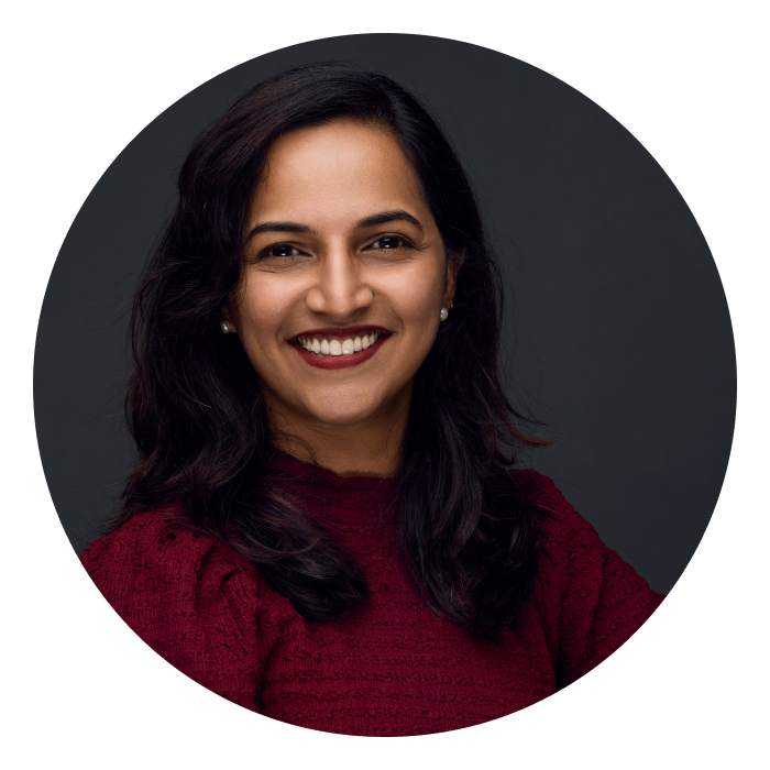 A headshot of Ramya, a middle-aged Indian woman with long dark hair, wearing a maroon top and a bright smile on her face.