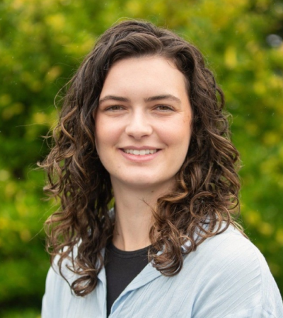 Siena is a middle aged Caucasian women with dark long curly hair and wearing a light gray shirt on top of black tshirt. She is smiling at the camera.