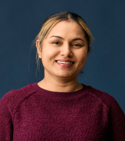 Camy is a middle-aged Indian woman with light brown skin and dark hair and dark brown eyes. She is wearing a maroon jumper and smiling at the camera.