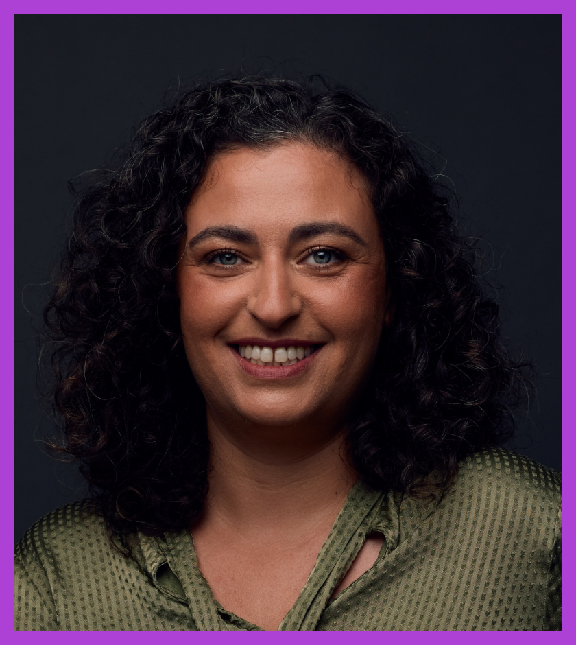Headshot of Maia - Maia is white with brown, shoulder-length curly hair and is smiling at the camera.