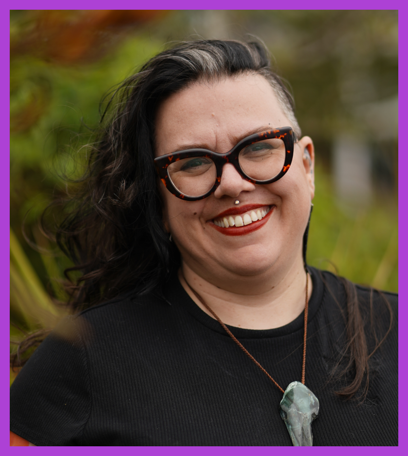 Headshot of Tamara, who has long brown hair, glasses, a big smile and has two full sleeve arm tattoos. 