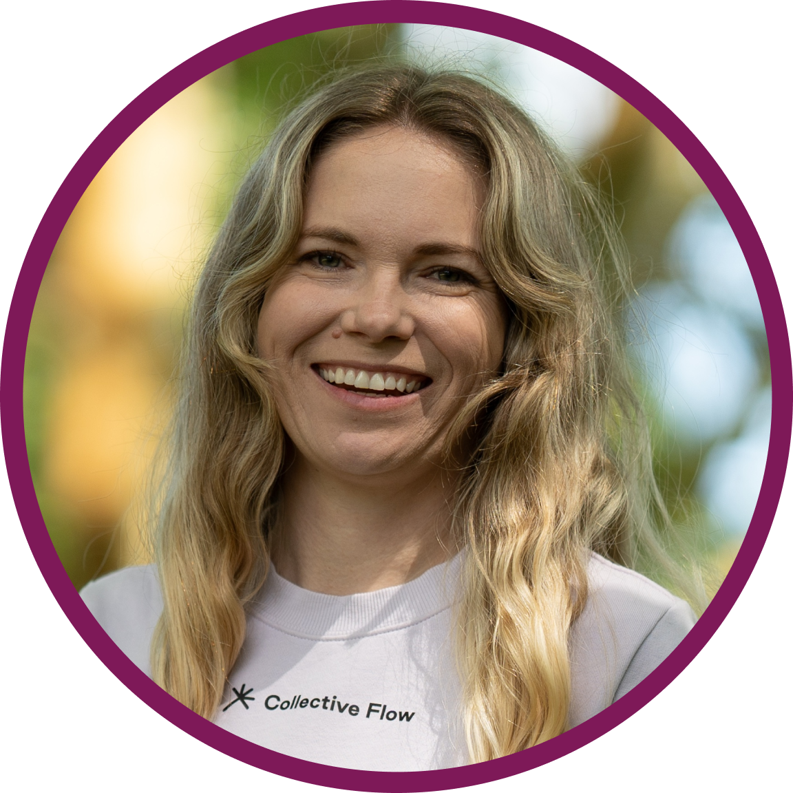 Bailey, a white woman with long wavy blonde hair, smiling at the camera. She’s standing in a leafy park under green trees, wearing a pale lilac Collective Flow t-shirt.