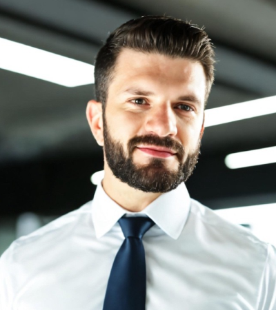 Tom has light skin, dark hair and beard, and is wearing a white formal shirt with a navy tie. 