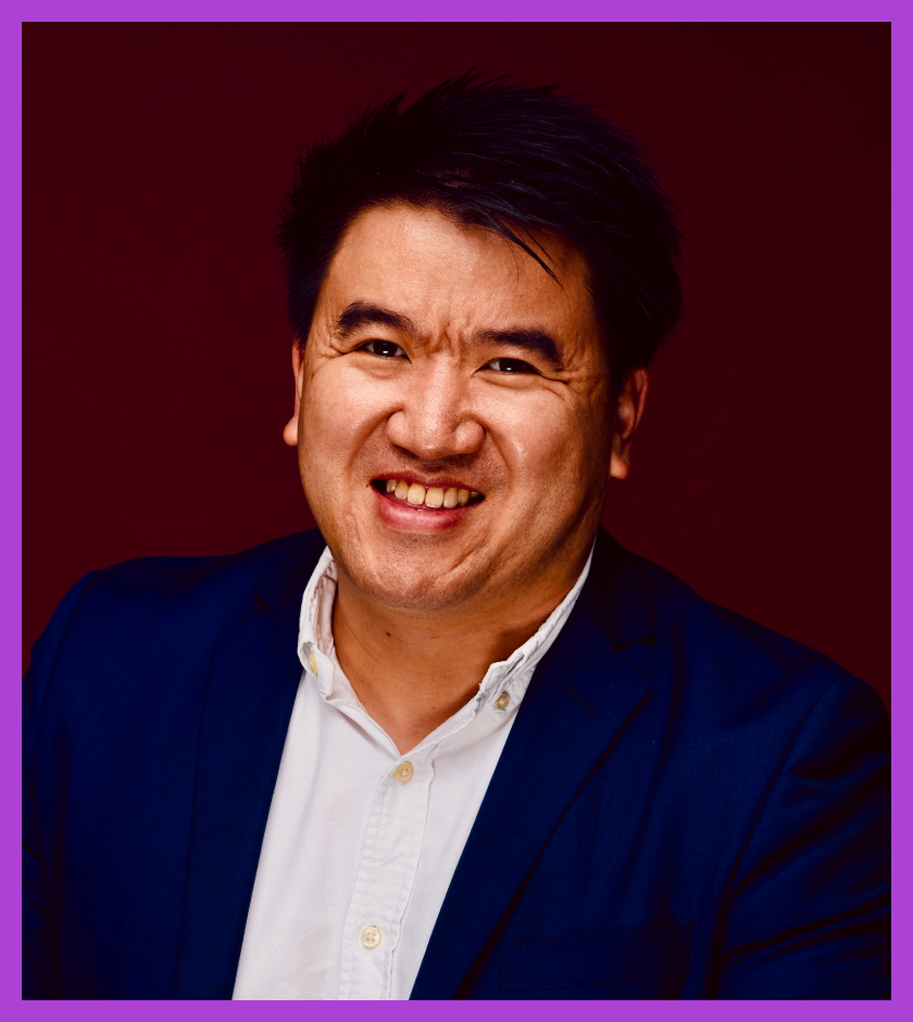 Headshot of Greg - an Asian male who is smiling, wearing a blue shirt and a pin collar suit jacket. 