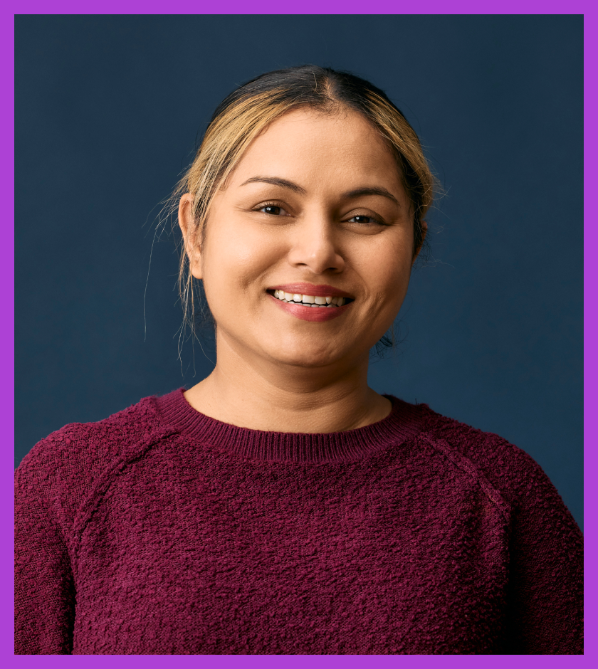 Camy is a middle-aged Indian woman with light brown skin and dark hair and dark brown eyes. She is wearing a maroon jumper and smiling at the camera. 