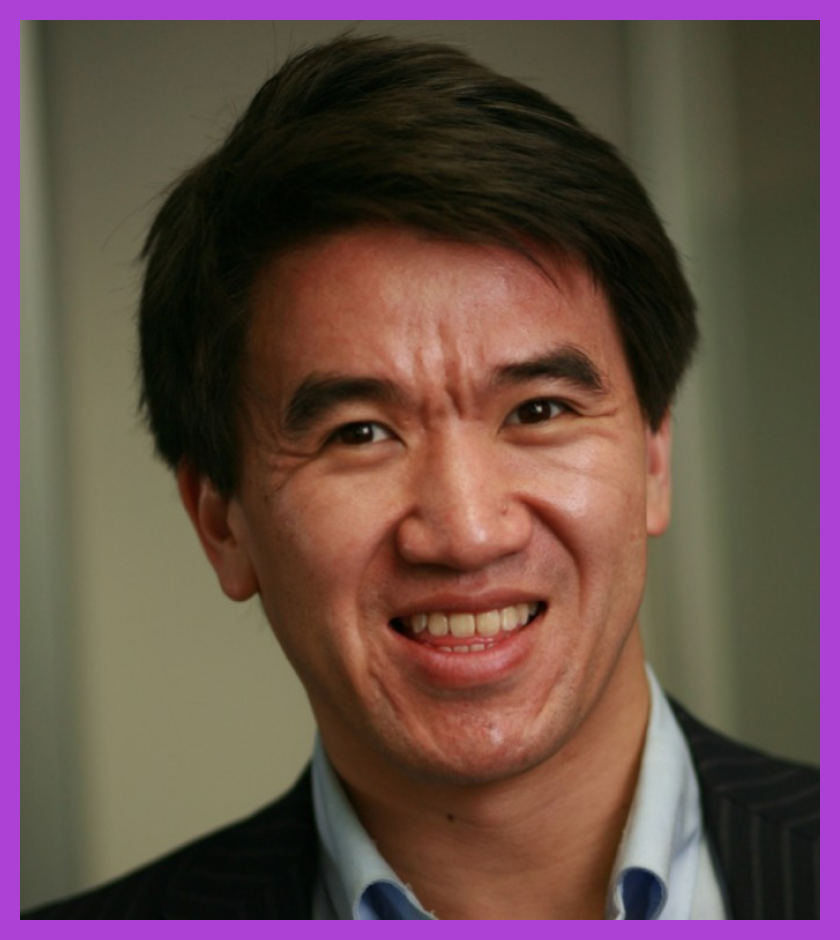 Headshot of Greg - an Asian male who is smiling, wearing a blue shirt and a pin collar suit jacket. 