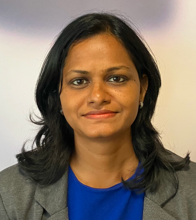 Surya is a woman with mid length dark hair, wearing a blue top and gray jacket. Surya is slightly smiling at the camera.