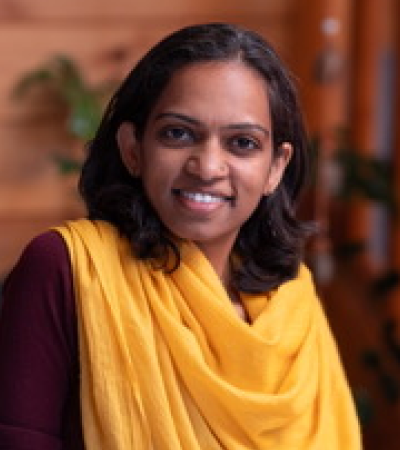 Sahana is a middle aged Indian women with dark hair wearing a maroon top and yellow shawl. She is smiling at the camera.