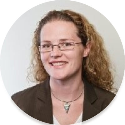 Ruth is a middel aged Caucasian woman with light brown curly hair, wearing glasses and a dark brown blazer, smiling at the camera.