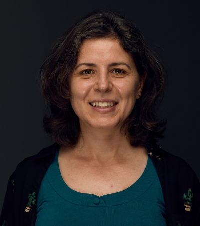 Lora is light skinned woman with light brown hair and wearing teal top and black cardigan, smiling at the camera.