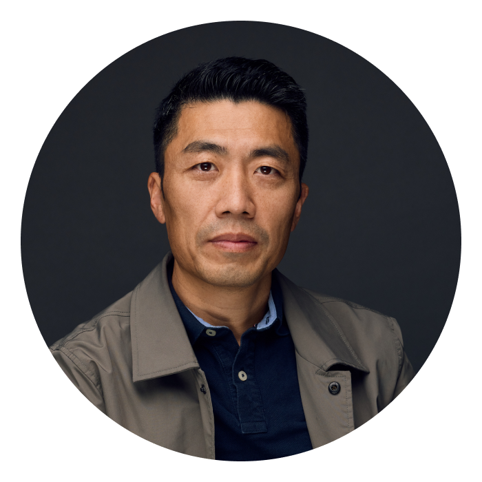 Headshot of Feilong - a middle-aged Chinese man with dark hair, and wearing a blue shirt and grey trench coat, looking at the camera.