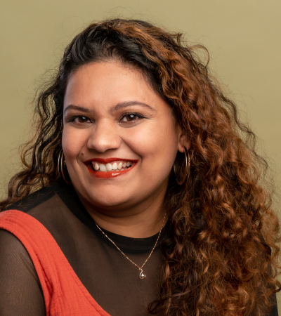 Headshot of Sneha - a woman with long curly hair, wearing a black tshirt and orange dress, smiling at the camera.