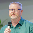 Toby is a middle-aged Caucasian man with light blonde and gray hair. He is wearing a green collar shirt and holding a mic in his hands. 