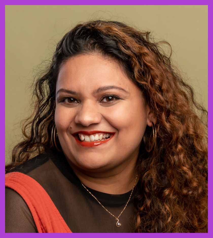 Headshot of Sneha - a woman with long curly hair, wearing a black tshirt and orange dress, smiling at the camera.