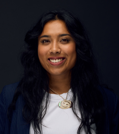 Aleisha is a young woman with brown skin and long dark hair. She is wearing a white t-shirt and a blue blazer, and smiling at the camera.