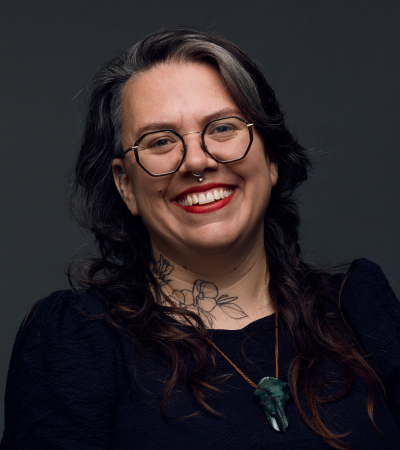 Headshot of Tamara, who has long brown hair, glasses, a big smile and has two full sleeve arm tattoos. 
