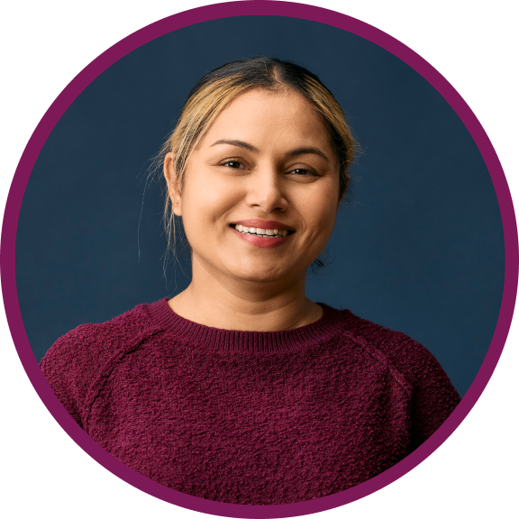 Camy is a middle-aged Indian woman with light brown skin and dark hair and dark brown eyes. She is wearing a maroon jumper and smiling at the camera. 