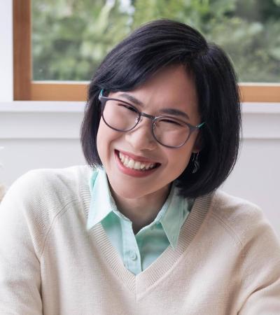 Prae is a middle-aged Thai woman with dark brown eyes and dark hair. She is wearing a white jumper and glasses, and smiling at the camera.
