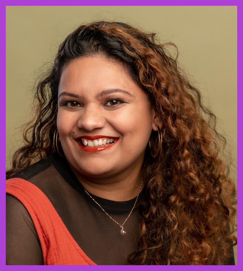 Headshot of Sneha - a woman with long curly hair, wearing a black tshirt and orange dress, smiling at the camera.