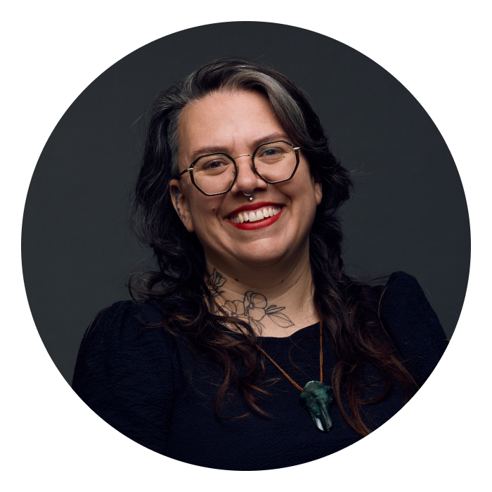 Headshot of Tamara, who has long brown hair, glasses, a big smile and has two full sleeve arm tattoos.