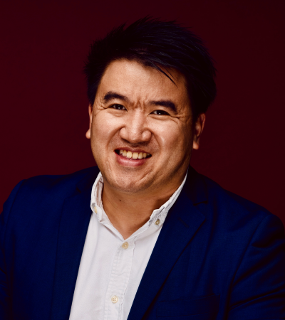 Headshot of Greg - an Asian male who is smiling, wearing a blue shirt and a pin collar suit jacket. 