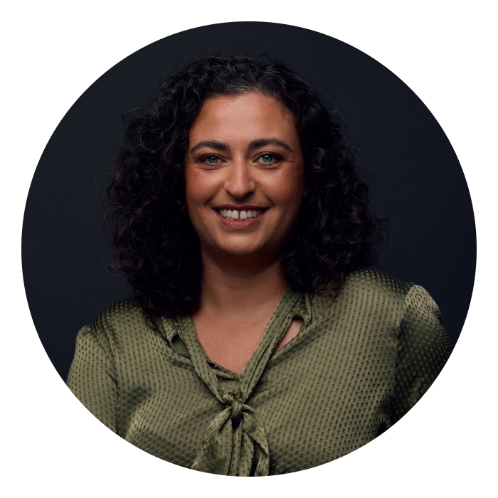 Headshot of Maia - Maia is white with brown, shoulder-length curly hair and is smiling at the camera.