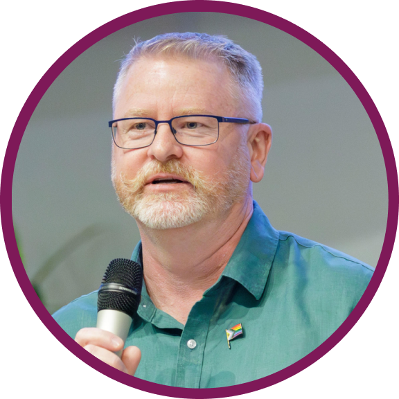 Toby is a middle-aged Caucasian man with light blonde and gray hair. He is wearing a green collar shirt and holding a mic in his hands. 