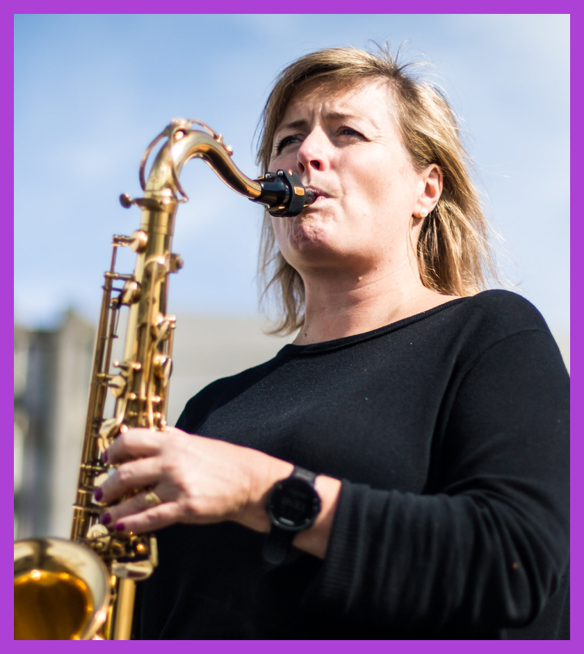 Rachel is a middle-aged Caucasian woman with blonde hair, wearing a black top and playing a saxophone.