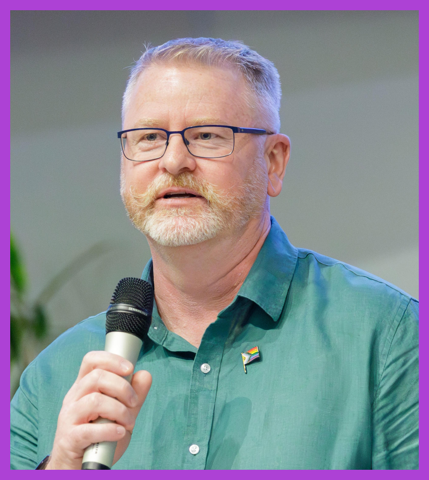 Toby is a middle-aged Caucasian man with light blonde and gray hair. He is wearing a green collar shirt and holding a mic in his hands.