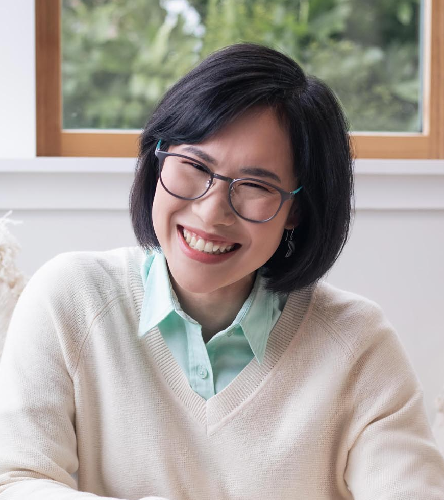 Prae is a middle-aged Thai woman with dark brown eyes and dark hair. She is wearing a white jumper and glasses, and smiling at the camera.