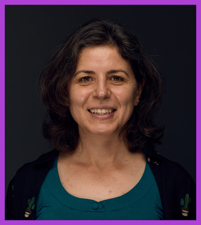 Lora is light skinned woman with light brown hair and wearing teal top and black cardigan, smiling at the camera.