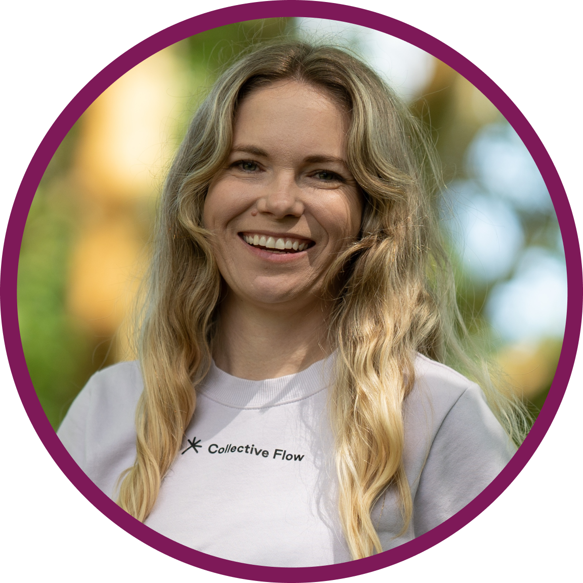 Bailey, a white woman with long wavy blonde hair, smiling at the camera. She’s standing in a leafy park under green trees, wearing a pale lilac Collective Flow t-shirt.