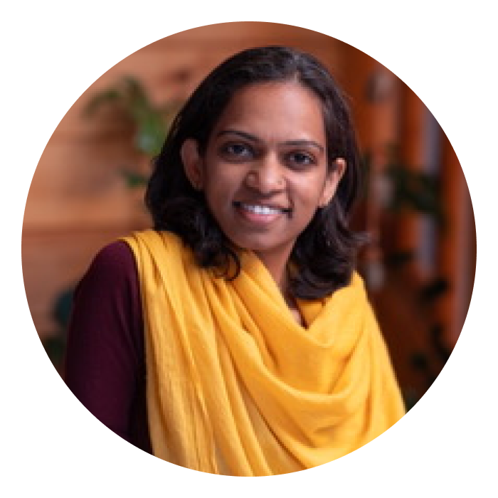 Sana is a middle aged Indian woman wearing a maroon top and a yellow wrap. She is smiling at the camera.