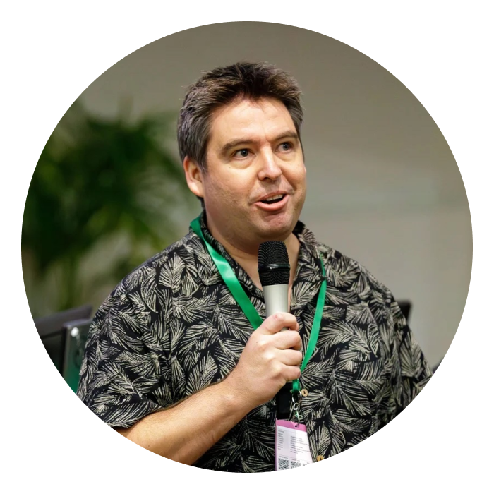 Aaron is a middle-aged Caucasian male, wearing a floral black shirt. He is holding a mic at his hand and looking at something.