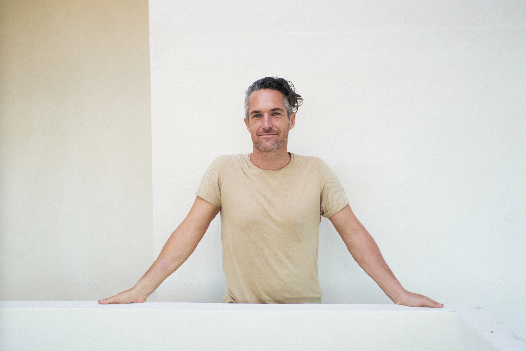 A man with gray hair and a beard, wearing a beige t-shirt, standing behind a white surface with his arms resting on it, smiling at the camera, against a plain white background.