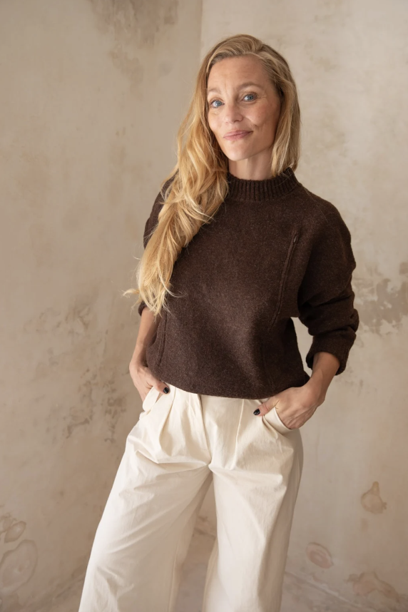 A woman with long blonde hair wearing a brown sweater and white pants, standing against a textured beige wall, looking at the camera with a slight smile. Fem, founder Liefs, retreat host, coach, facilitator. Rancho Verde.