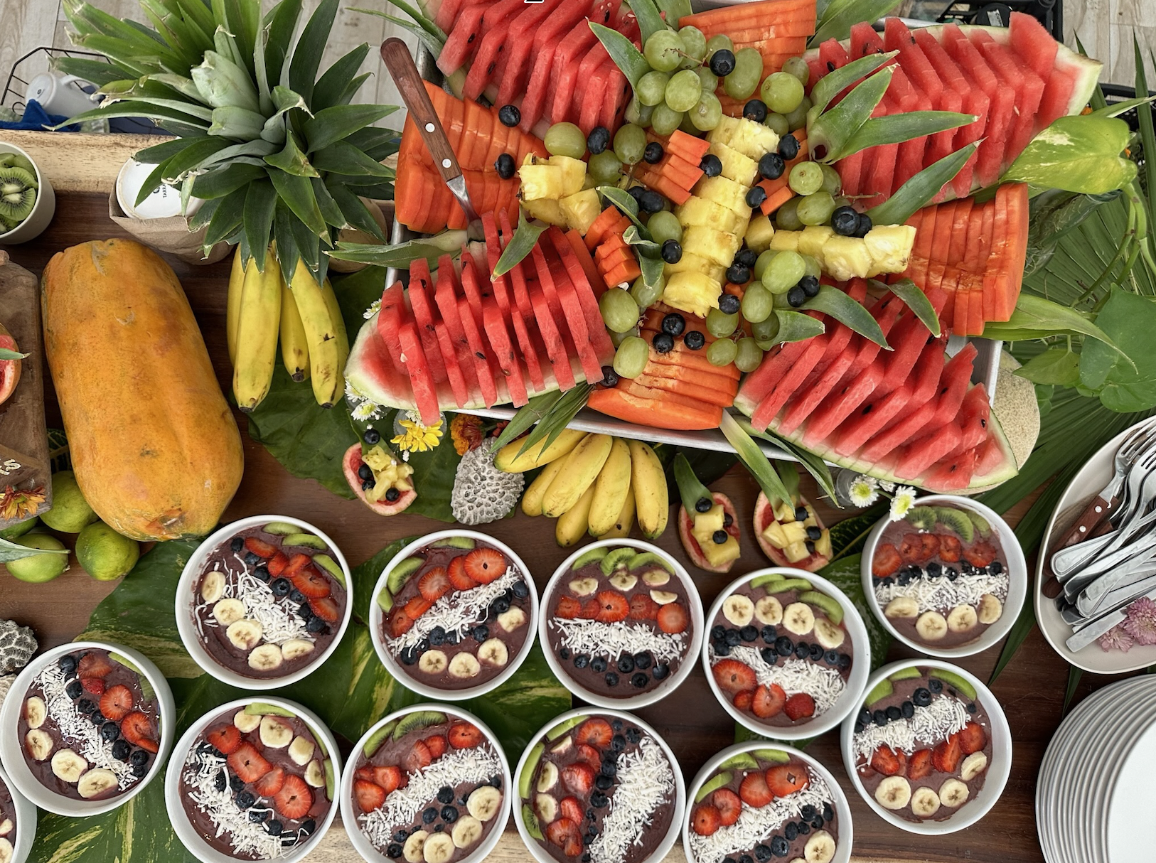 A colorful fruit display featuring sliced watermelons, pineapples, grapes, bananas, and assorted berries arranged on a table with bowls of fruit and a large mango.