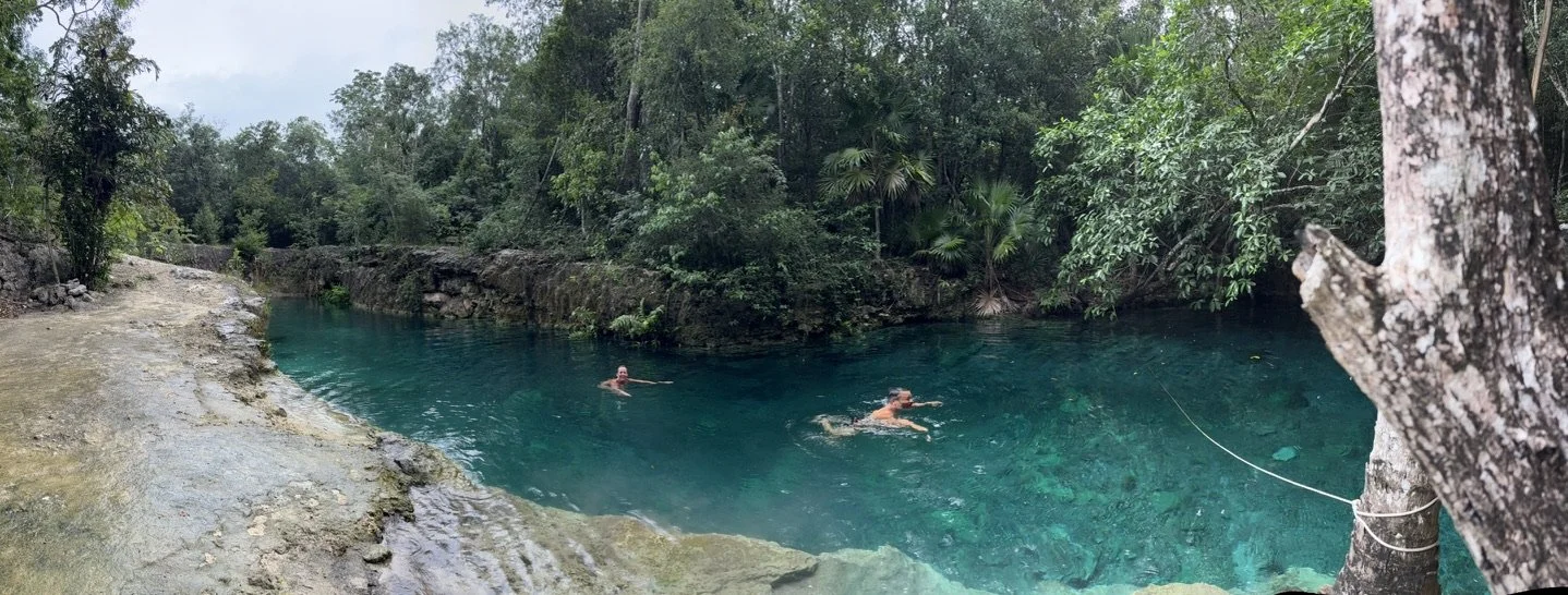 Two people swimming in a clear, blue-green river surrounded by dense trees and rocky banks.