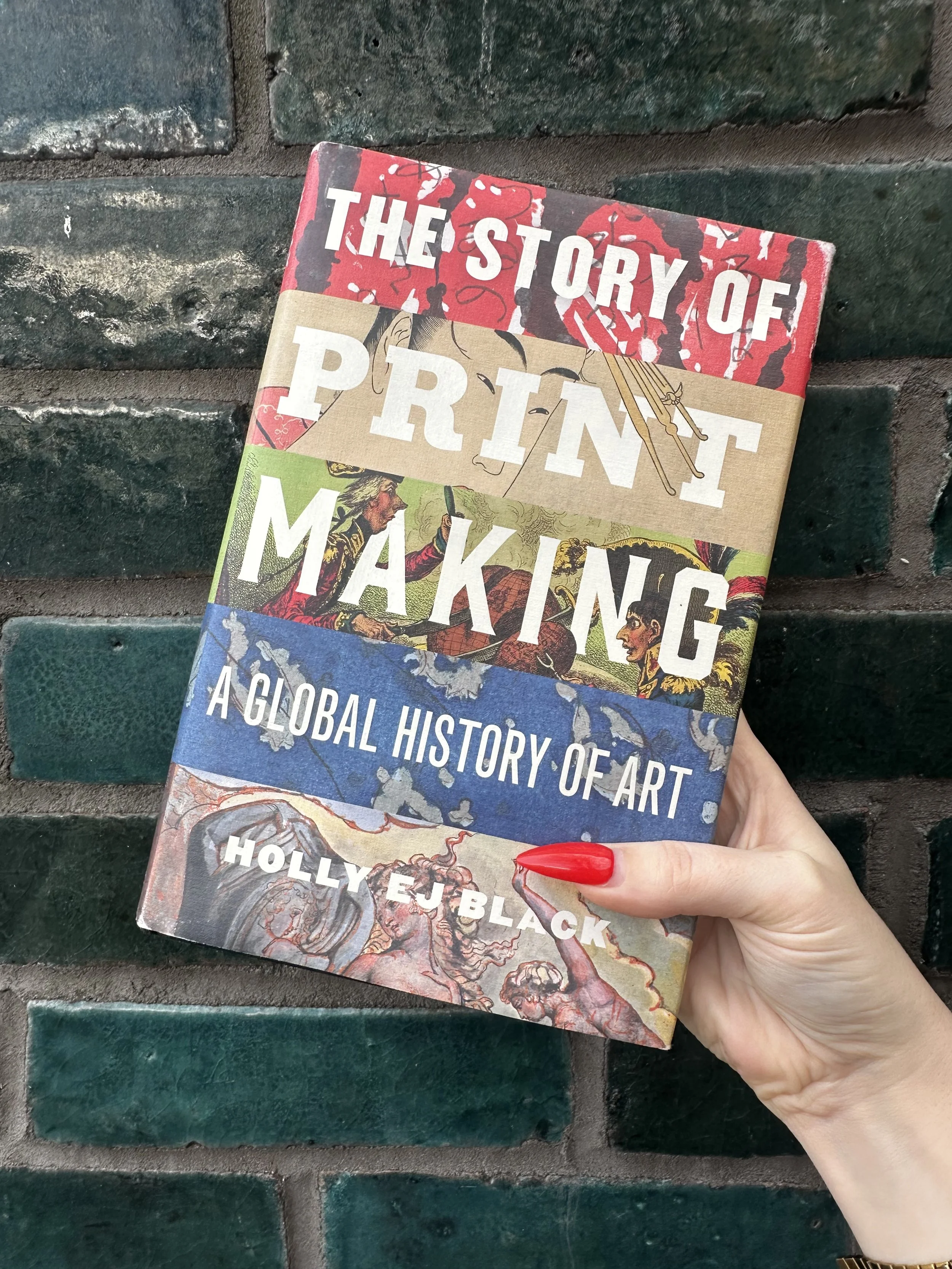 hand holding the book 'the story of printmaking' in front of a green brick wall