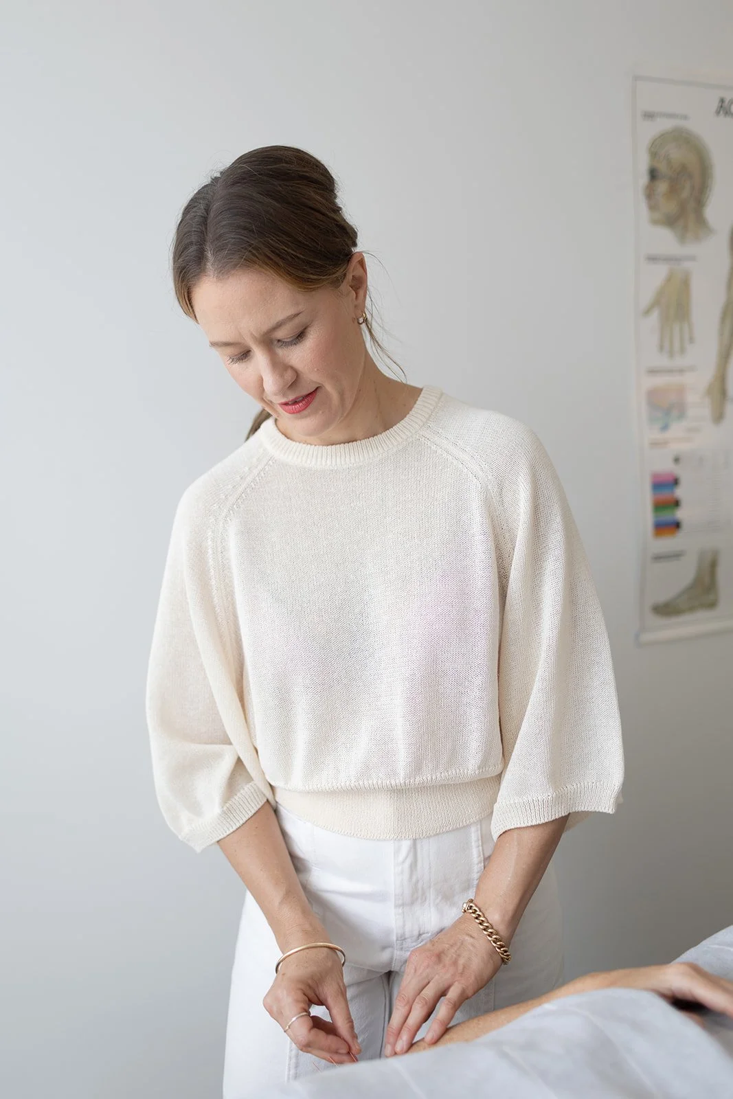 A woman in a white sweater and white pants appears to be performing acupuncture on a patient who is lying down, with an acupuncture meridian chart in the background.