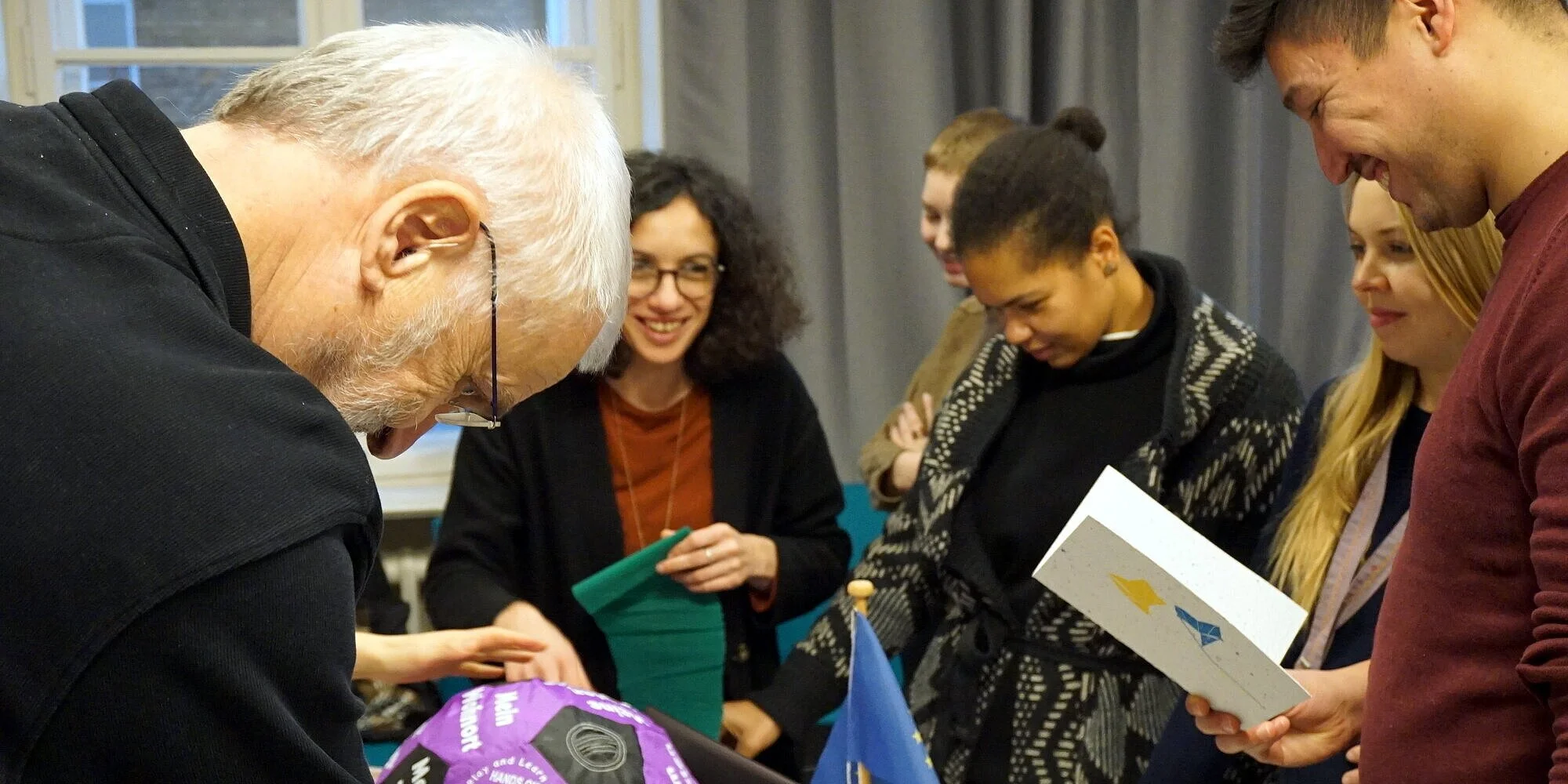 Im Vordergrund ein älterer Mann mit Brille; im Hintergrund eine Gruppe fröhlicher, junger Leute. Gemeinsam schauen sich alle etwas an.