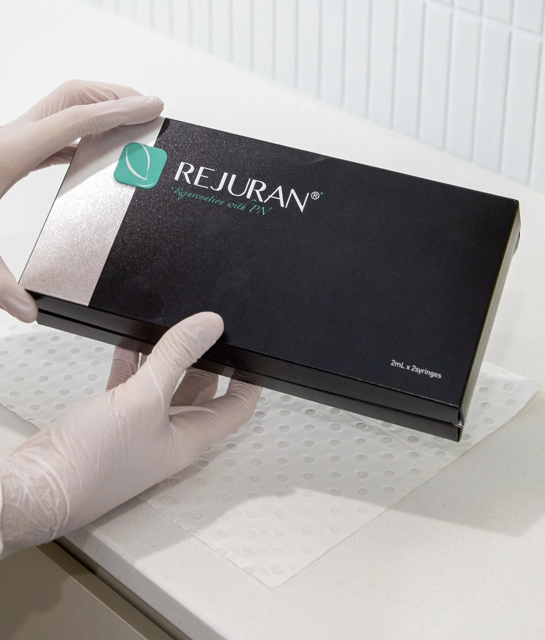 A person wearing gloves holding a black box labeled Rejuvan Quotation with PV, containing 2ml x 25 syringes, on a white surface with a textured paper underneath.