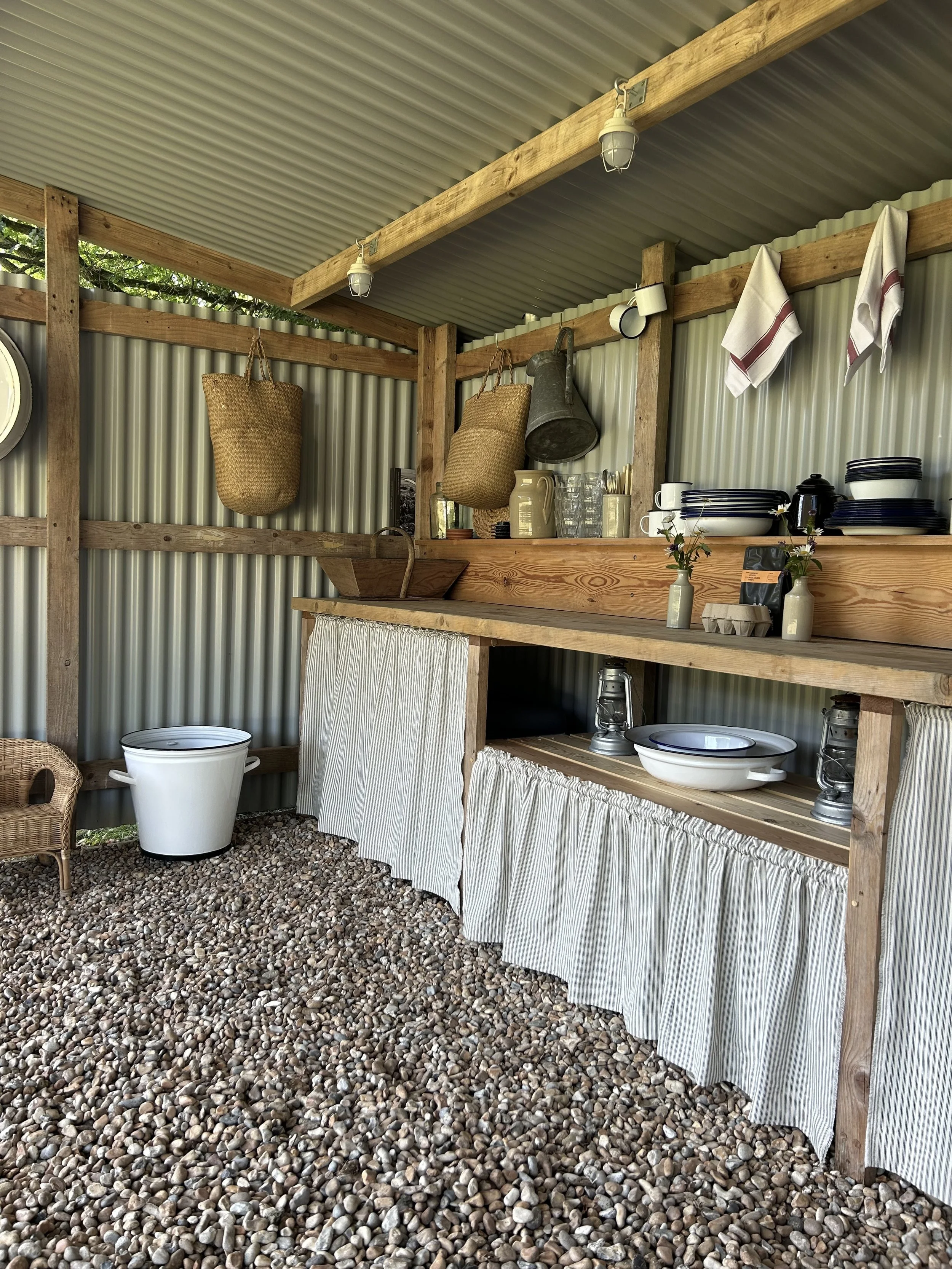 Field kitchen details - corrugated metal roof, wooden beams, and a gravel floor. There are hanging woven baskets, white towels with red stripes, a collection of dishes and glasses, a small potted plant, a vintage lantern, and a white enamel bucket.