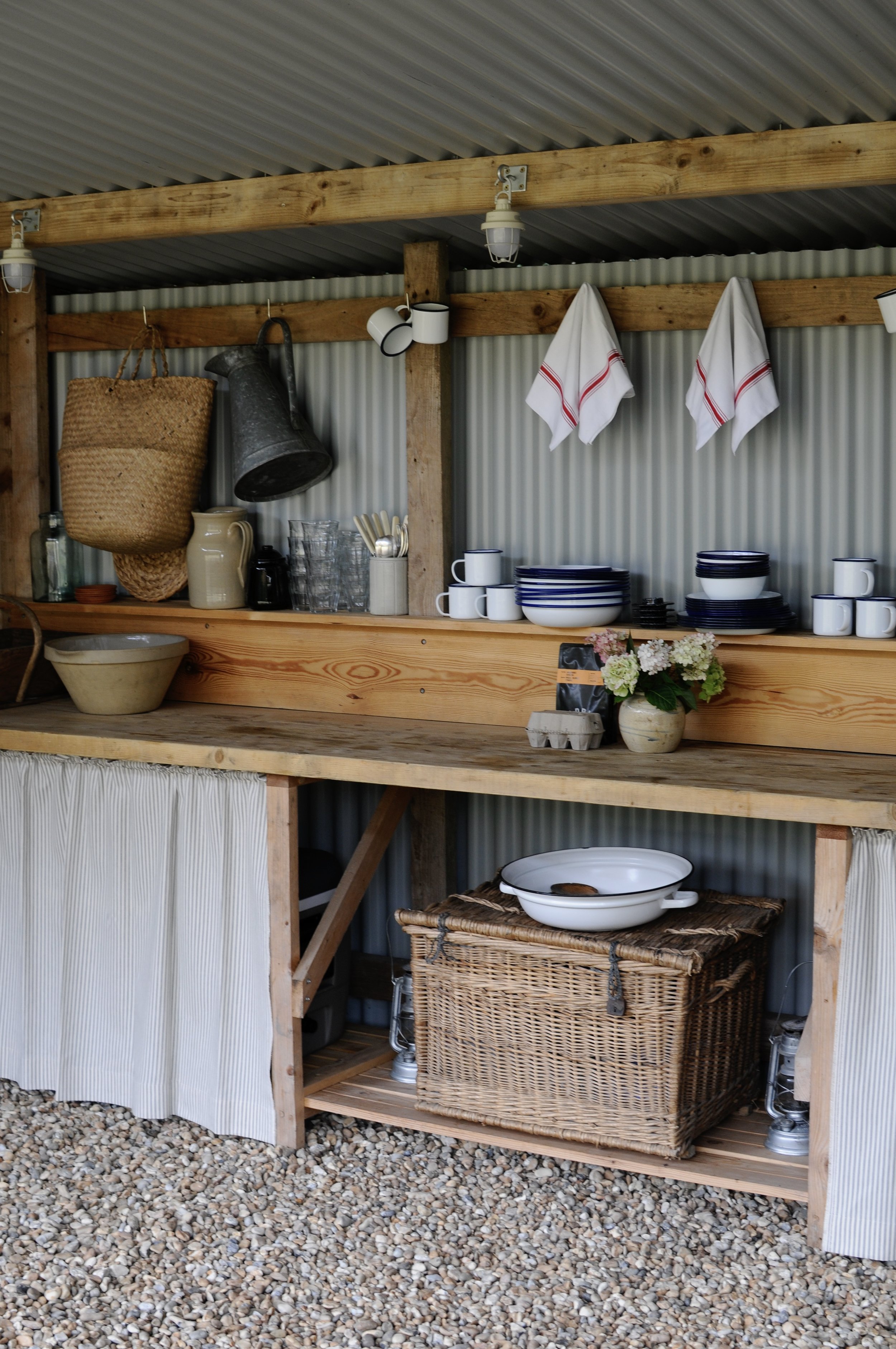 Part of an outdoor kitchen or patio area with a counter, wooden beams, hanging towels, cups, plates, a basket, and a flowerpot, with a gravel ground.