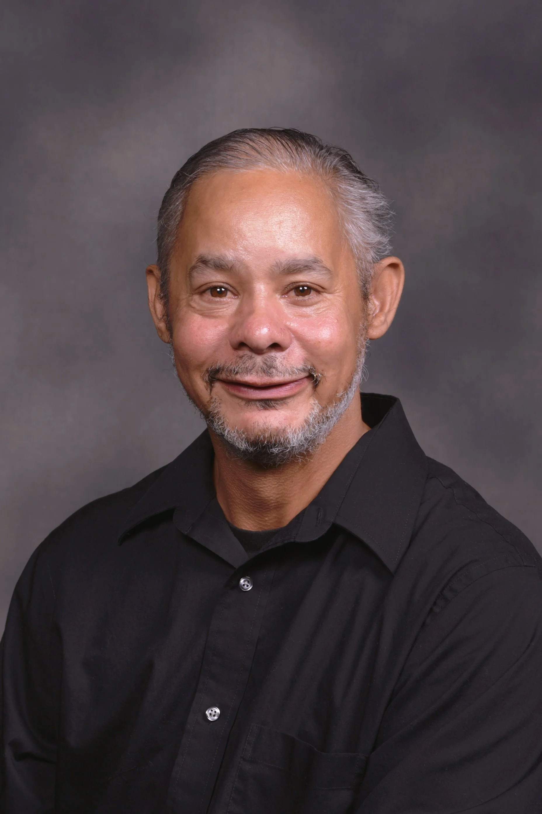 A man with gray hair and a goatee, wearing a dark T-shirt, smiling indoors with wooden paneling and text in the background.