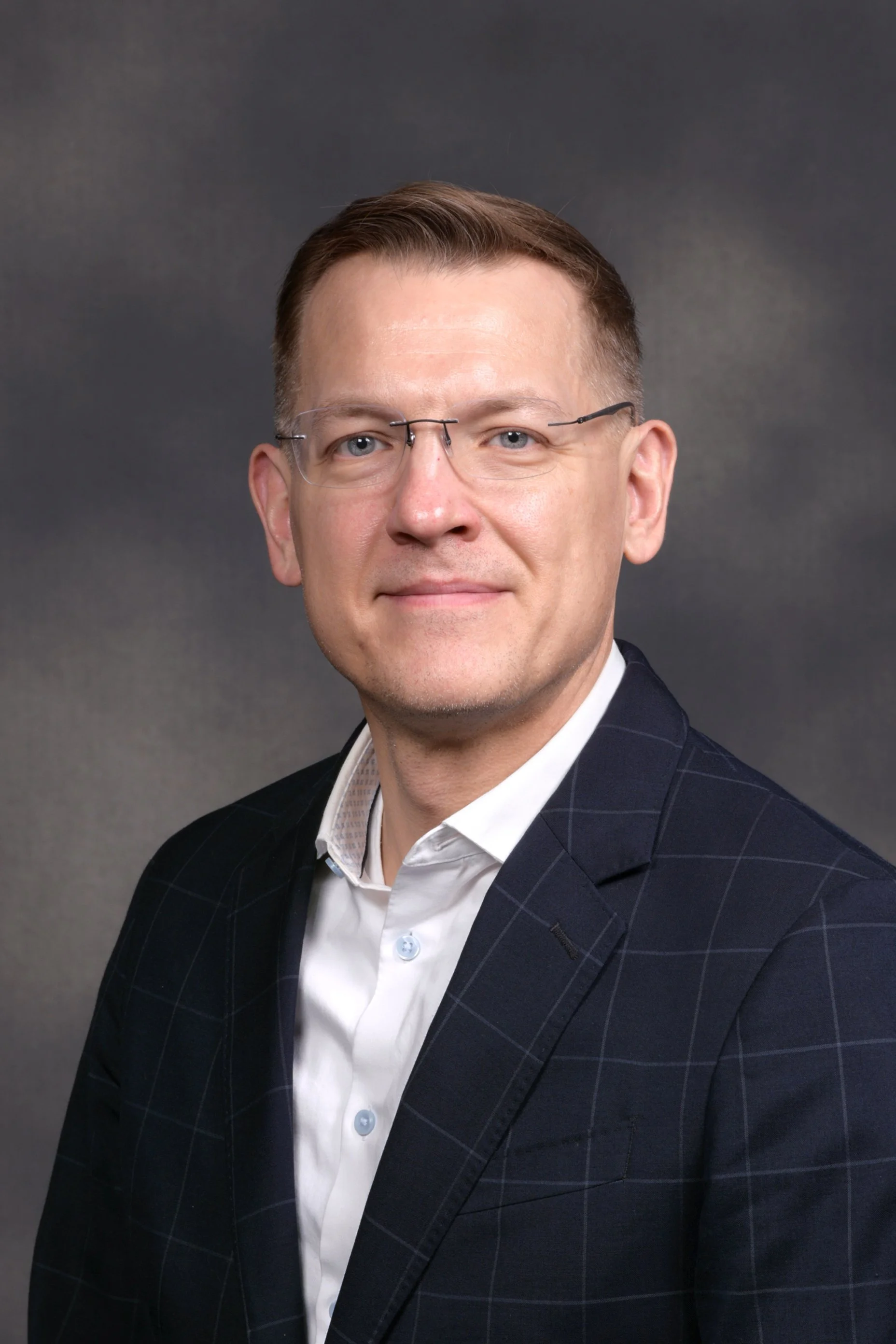 A smiling man with glasses, short brown hair, wearing a black suit and tie, against a plain light background.