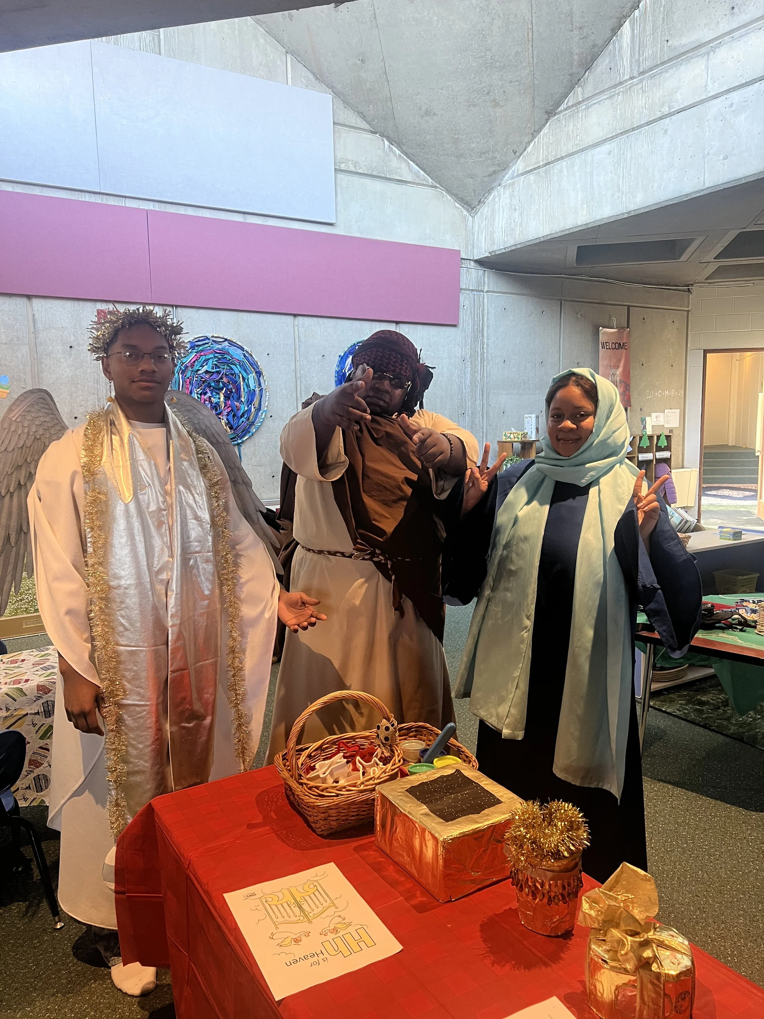 Three people dressed in costumes standing behind a table decorated for Christmas, with festive items like a gift box, basket, and sign on the table.