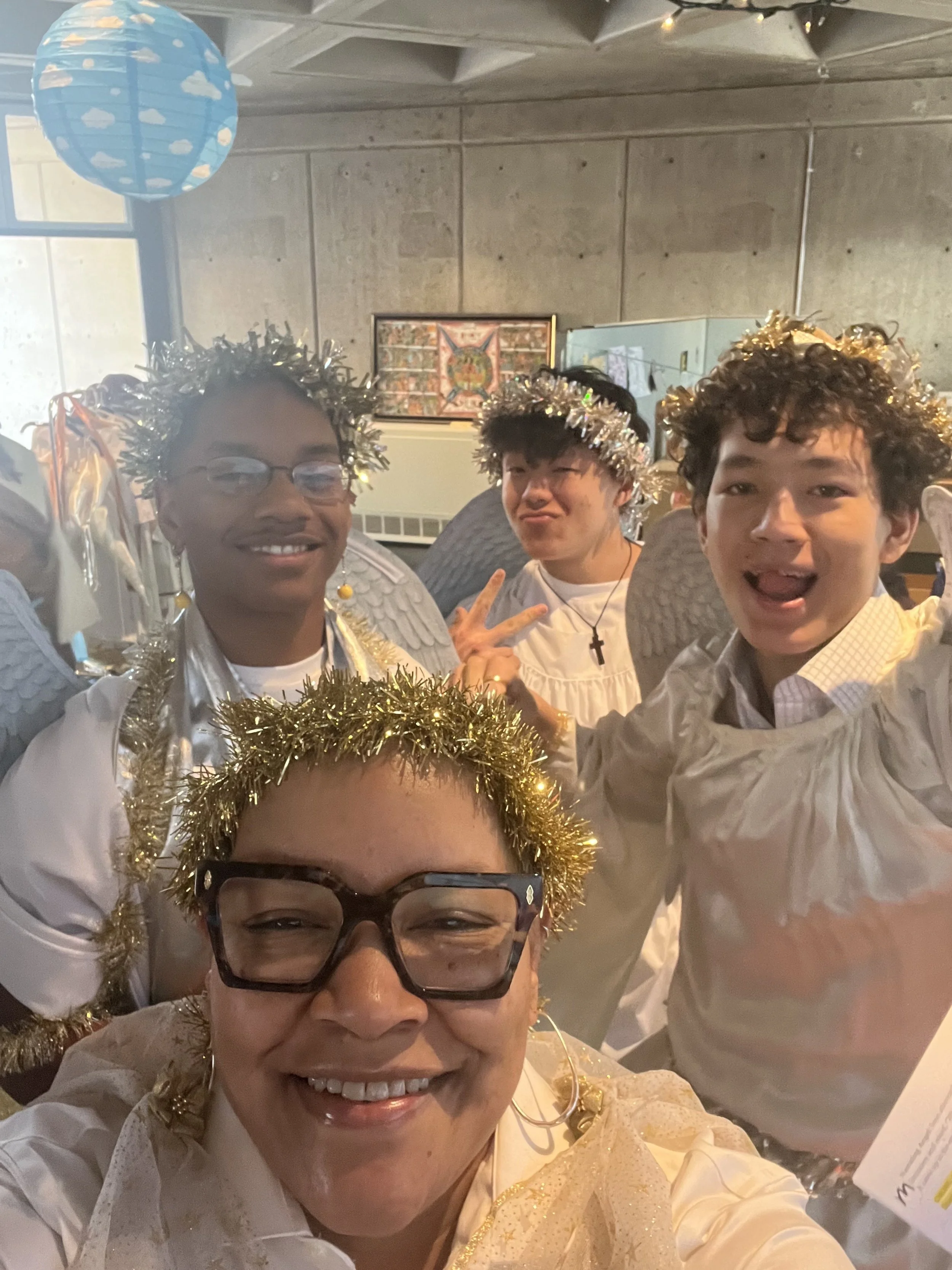 A group of five people dressed in angel costumes with tinsel halos and wings, smiling and posing for a photo indoors.