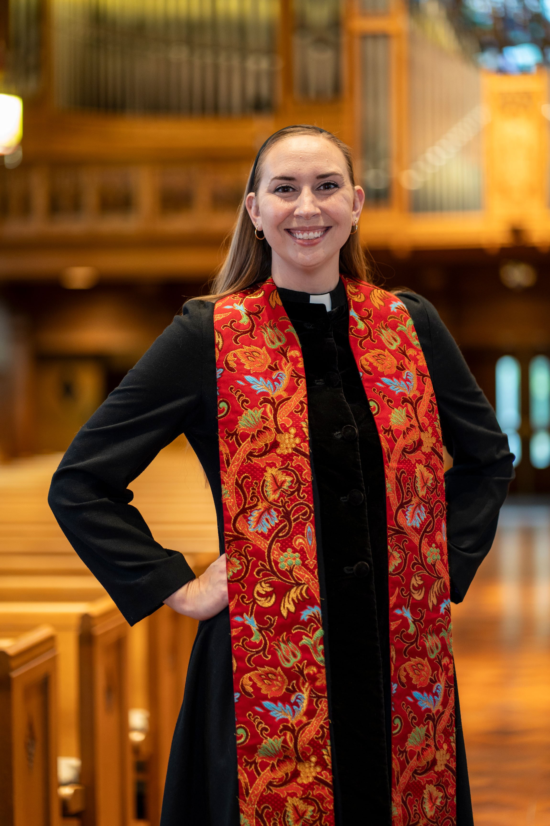 Rev. Garry smiling wearing a clerical collar, black dress, and a red embroidered stole standing inside a church with wooden pews and stained glass windows in the background.